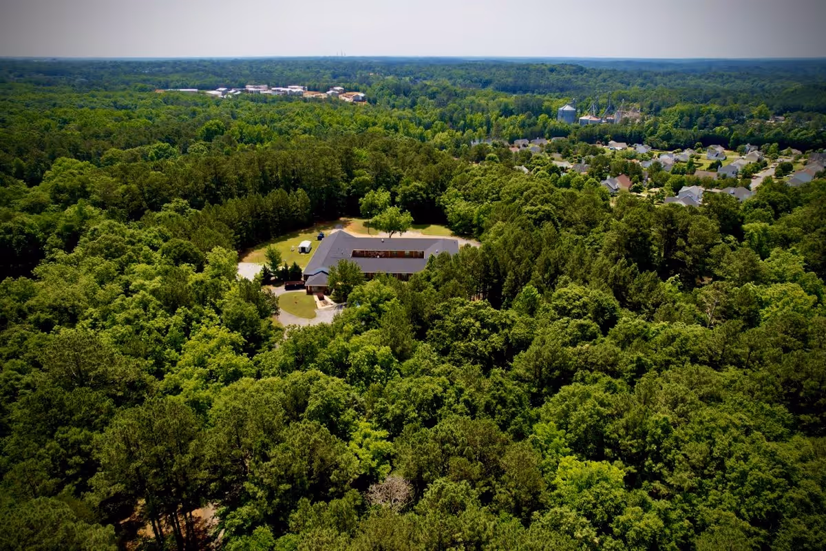 Aerial view of a building surrounded by dense green forest with a residential neighborhood visible in the distance under a clear sky.