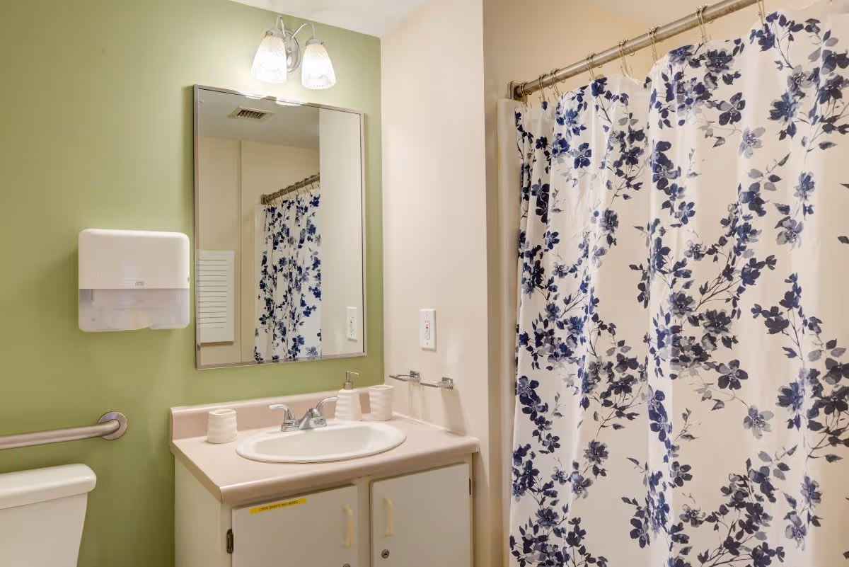 A bathroom with a green and beige wall, a white sink with a countertop, a mirror above the sink, a light fixture with two bulbs, a toilet with a grab bar, a paper towel dispenser, and a shower with a white curtain featuring a blue floral pattern.