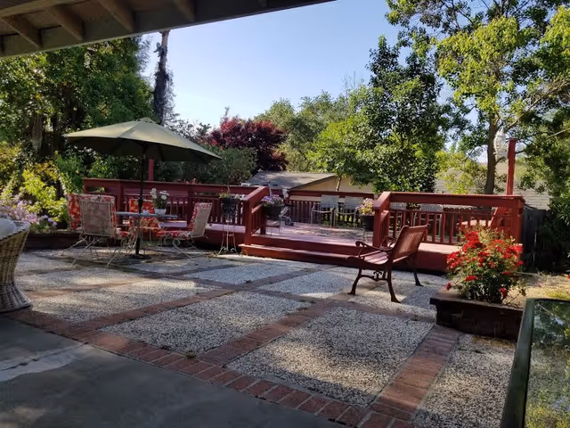Outdoor patio area with a red wooden deck, several chairs, a table with a large umbrella, and various plants and trees surrounding the space under a clear blue sky.