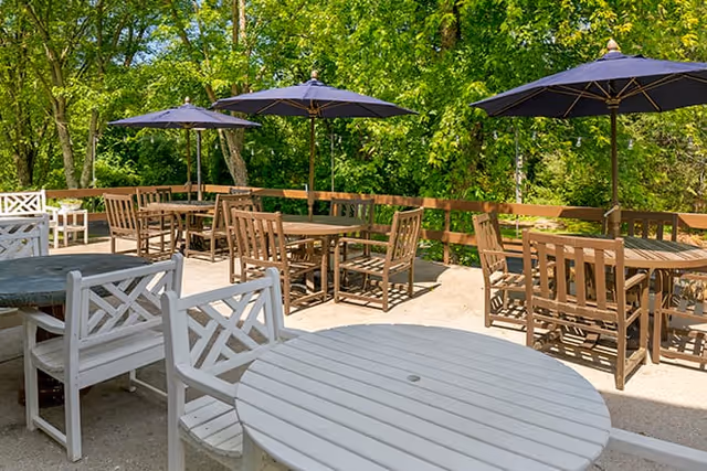 Outdoor patio area with multiple wooden tables and chairs, some tables shaded by dark blue umbrellas, surrounded by green trees and foliage.