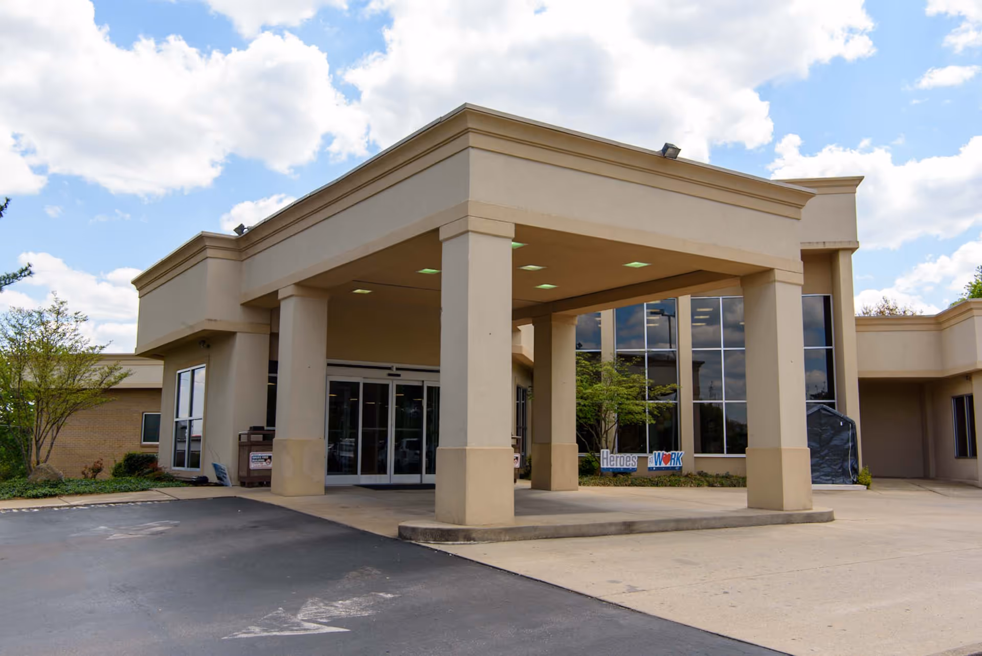 Exterior view of the entrance to Bethany Center for Rehabilitation and Healing, featuring a covered drop-off area with large pillars, glass doors, and windows reflecting the sky and surrounding greenery under a partly cloudy sky.