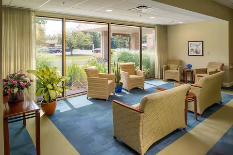A bright and cozy sitting area with large floor-to-ceiling windows letting in natural light. The room features several cushioned armchairs and sofas arranged around small wooden tables. There are potted plants and flowers placed near the windows and on a side table, with a framed artwork hanging on the wall. Outside the windows, greenery and a parking lot are visible.