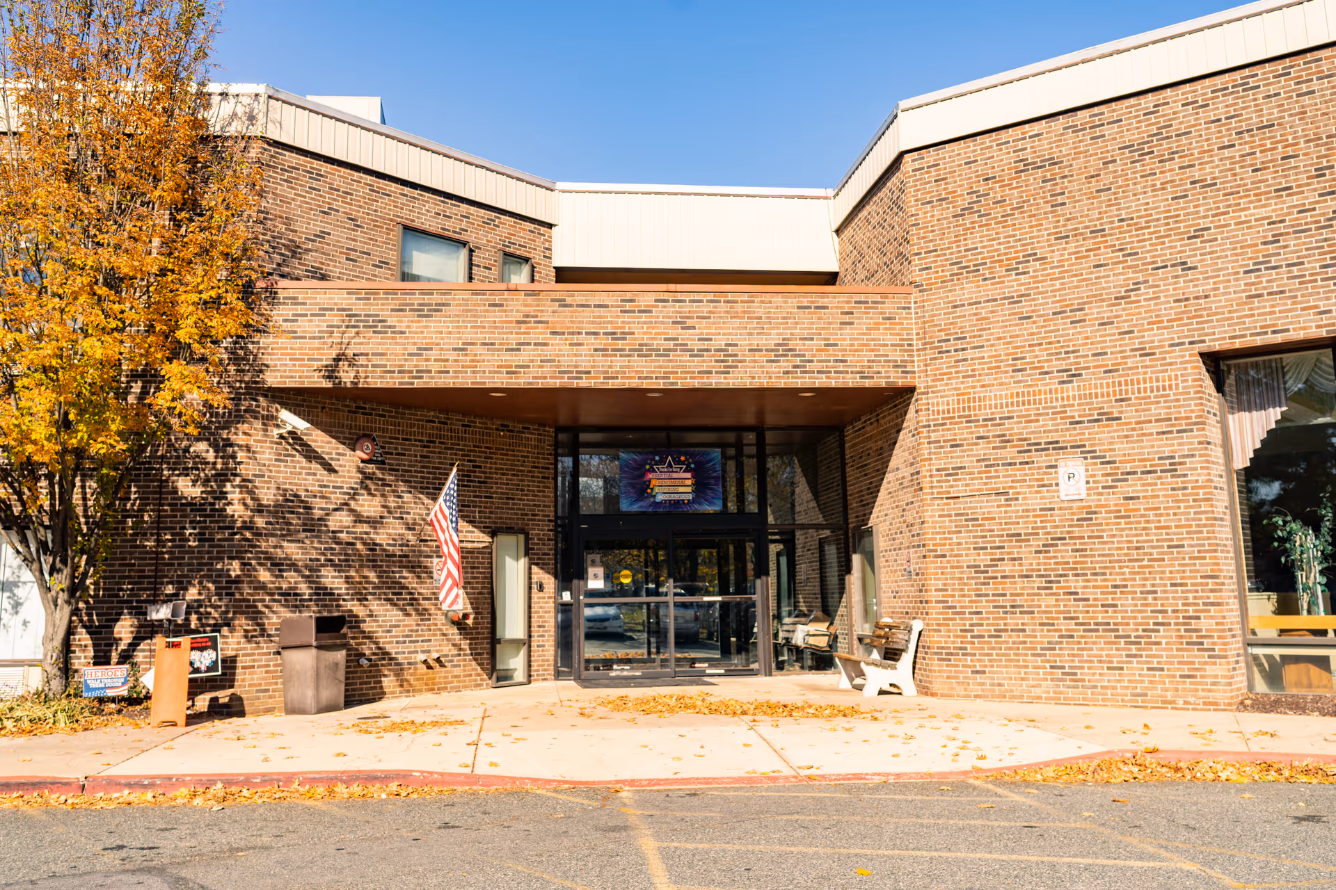 Exterior front entrance of a brick building with a covered entryway, an American flag mounted on the wall, a bench, and a tree with autumn-colored leaves on the left side.