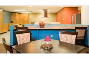 Interior view of a dining area with a table and chairs in the foreground, decorated with a small centerpiece and a pink lamp. In the background, there is a kitchen area with a countertop, cabinets, a refrigerator, and a range hood. The walls are painted in warm tones including orange and beige.
