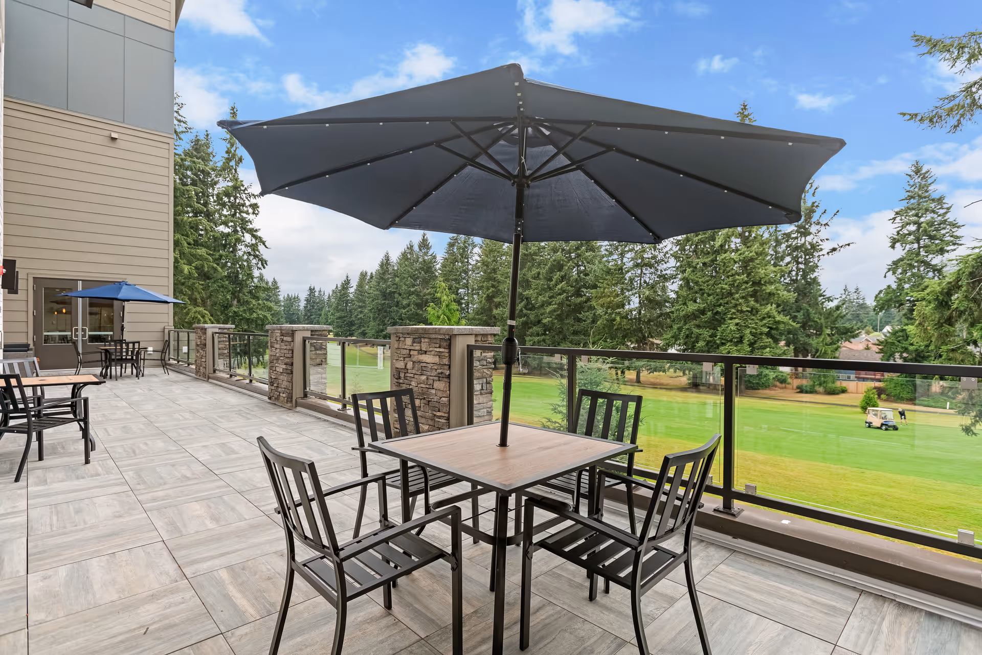 Outdoor patio area with tables and chairs, each table shaded by a large umbrella. The patio overlooks a green lawn and trees, with a golf cart visible in the distance. The building exterior is visible on the left side.