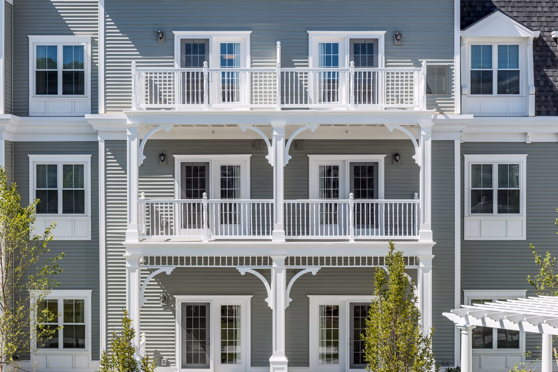 Exterior view of a senior living facility building with gray siding and white trim. The building features two levels of balconies with white railings and decorative supports. Several windows and glass doors open onto the balconies. Small trees and shrubs are visible in front of the building.