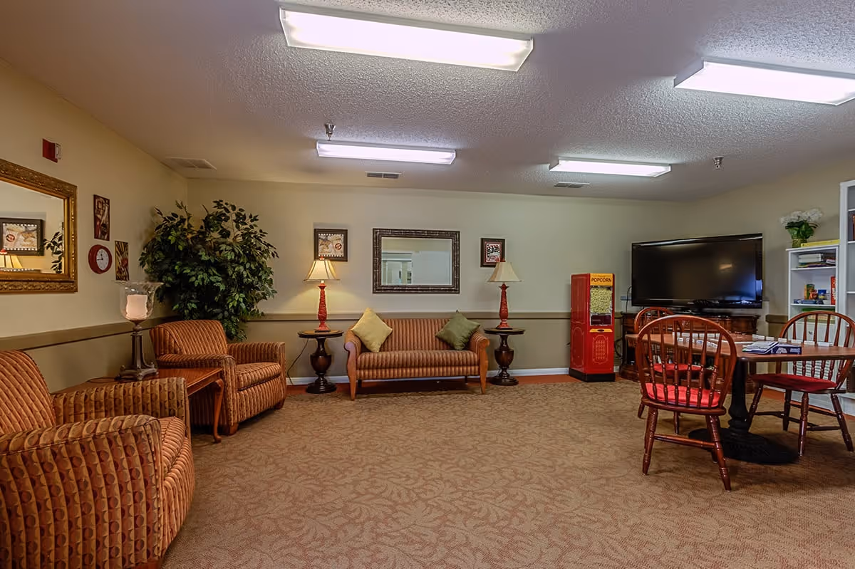 A cozy senior living lounge with upholstered chairs and a sofa, lamps, a TV, a small table with chairs, and a popcorn machine.