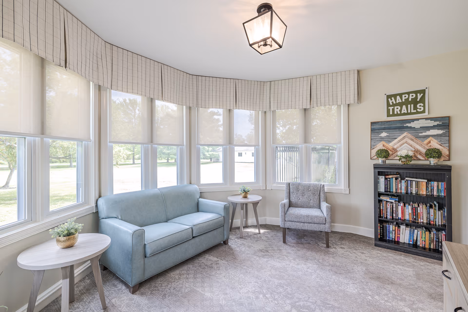 A bright and cozy sitting area with a light blue sofa, a gray armchair, two small round wooden side tables with potted plants, a black bookshelf filled with books, and large windows with light-colored blinds and valances. A wall decoration above the bookshelf reads 'HAPPY TRAILS'.