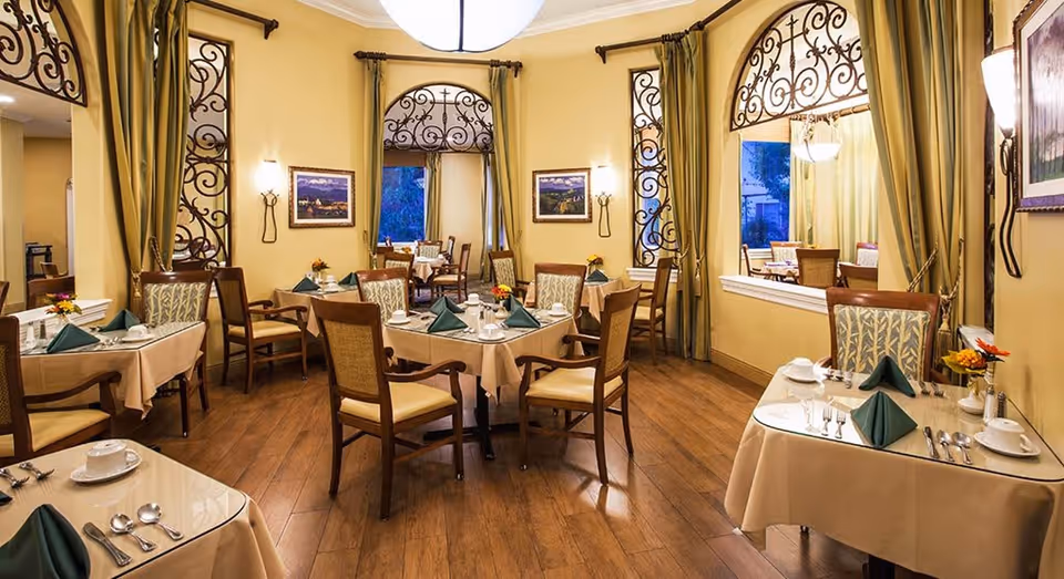 A warmly lit dining room with several tables covered in beige tablecloths, each set with green folded napkins, white cups, saucers, and silverware. The room features wooden chairs with cushioned seats, large windows with decorative wrought iron grills, and yellow walls adorned with framed artwork and wall sconces. The floor is wooden, and there are curtains hanging beside the windows.