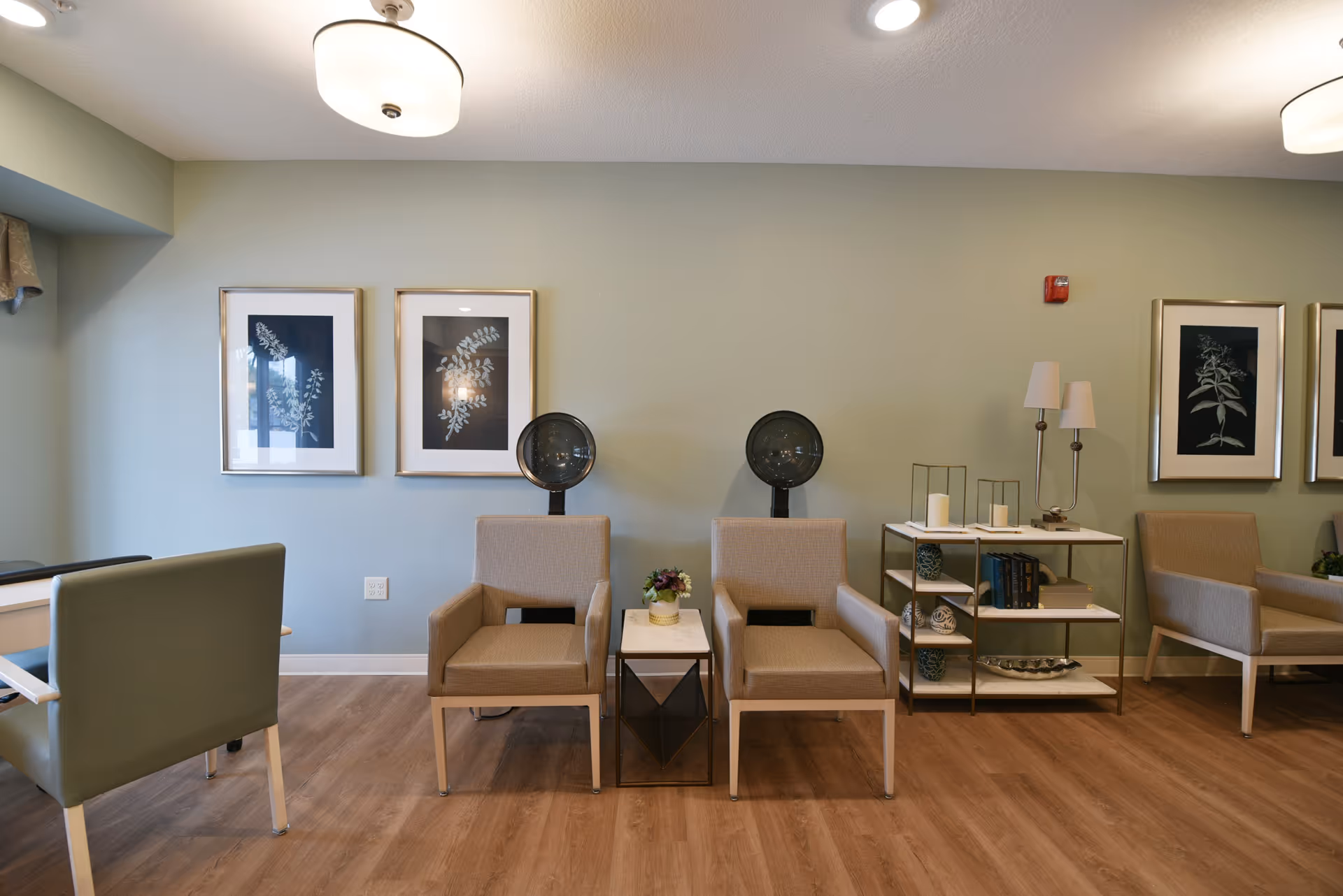 Interior view of a salon area in a senior living facility with two beige salon chairs under hair dryers, a small table with a flower arrangement between them, a shelving unit with decorative items and books, and framed botanical artwork on a light green wall.