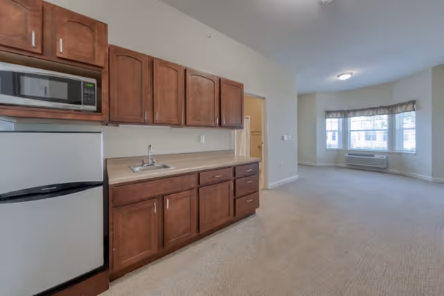 Interior view of a senior living facility apartment showing a small kitchen area with wooden cabinets, a microwave, a mini refrigerator, and a sink. The kitchen opens into a carpeted living space with a large window and an air conditioning unit below it.