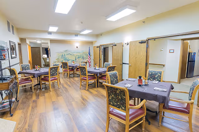 A dining room with several tables covered in dark tablecloths, each set with napkins and utensils. The room has wooden floors, wooden chairs with patterned upholstery, and beige walls. There is a mural on the far wall and an American flag near the back. The room is well-lit with ceiling lights and has open doorways leading to other areas.