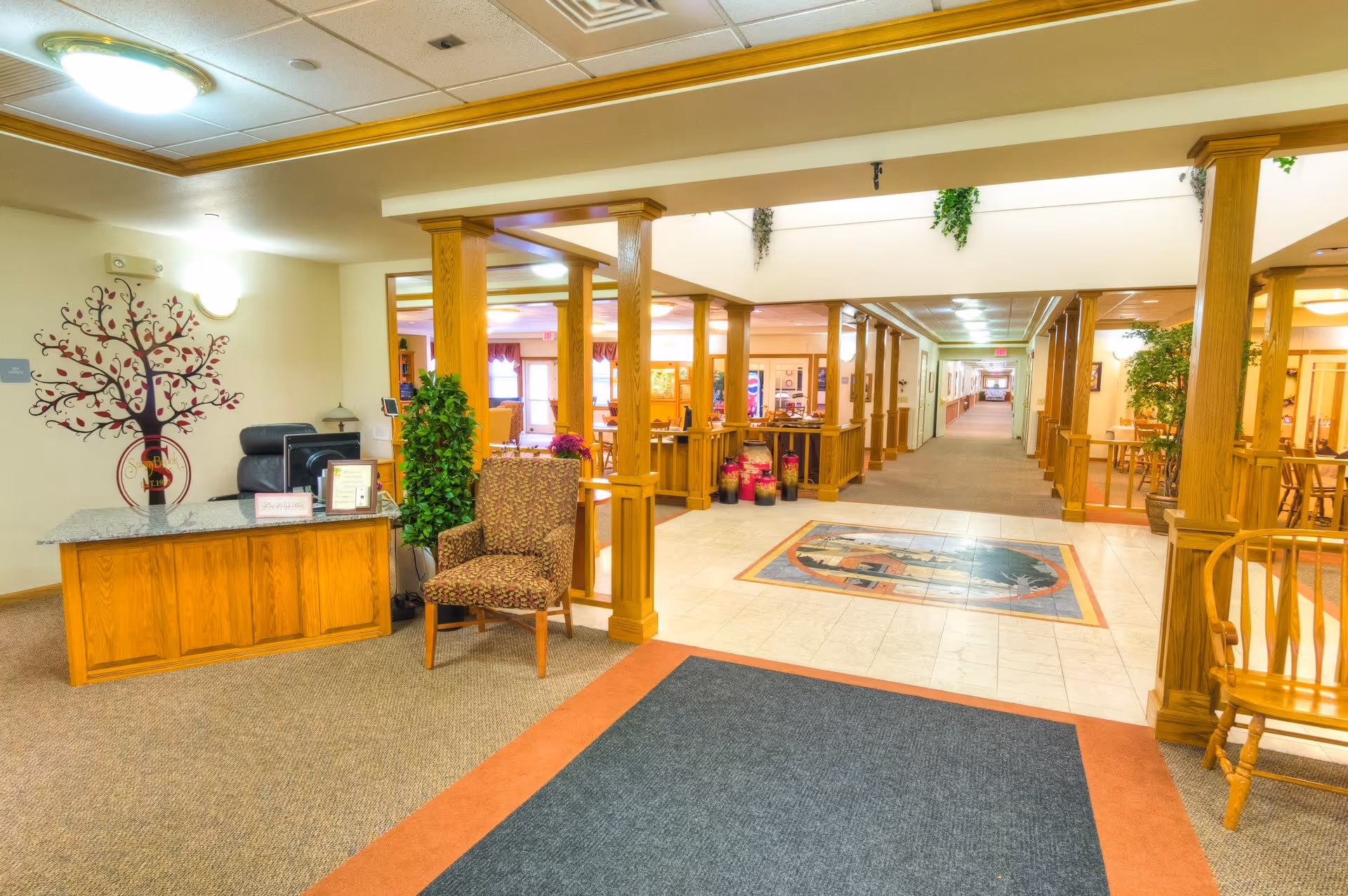 Reception area of a senior living facility with a wooden front desk, a patterned armchair, decorative plants, and a hallway leading to other rooms. The space features wooden pillars, a tiled floor section with a decorative design, and carpeted areas.