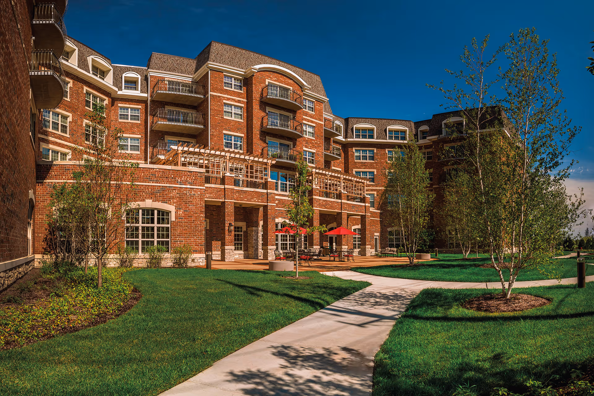 Exterior view of a multi-story brick building with balconies and large windows, surrounded by a well-maintained lawn, young trees, and a paved walkway leading to a patio area with red umbrellas and outdoor seating under a clear blue sky.