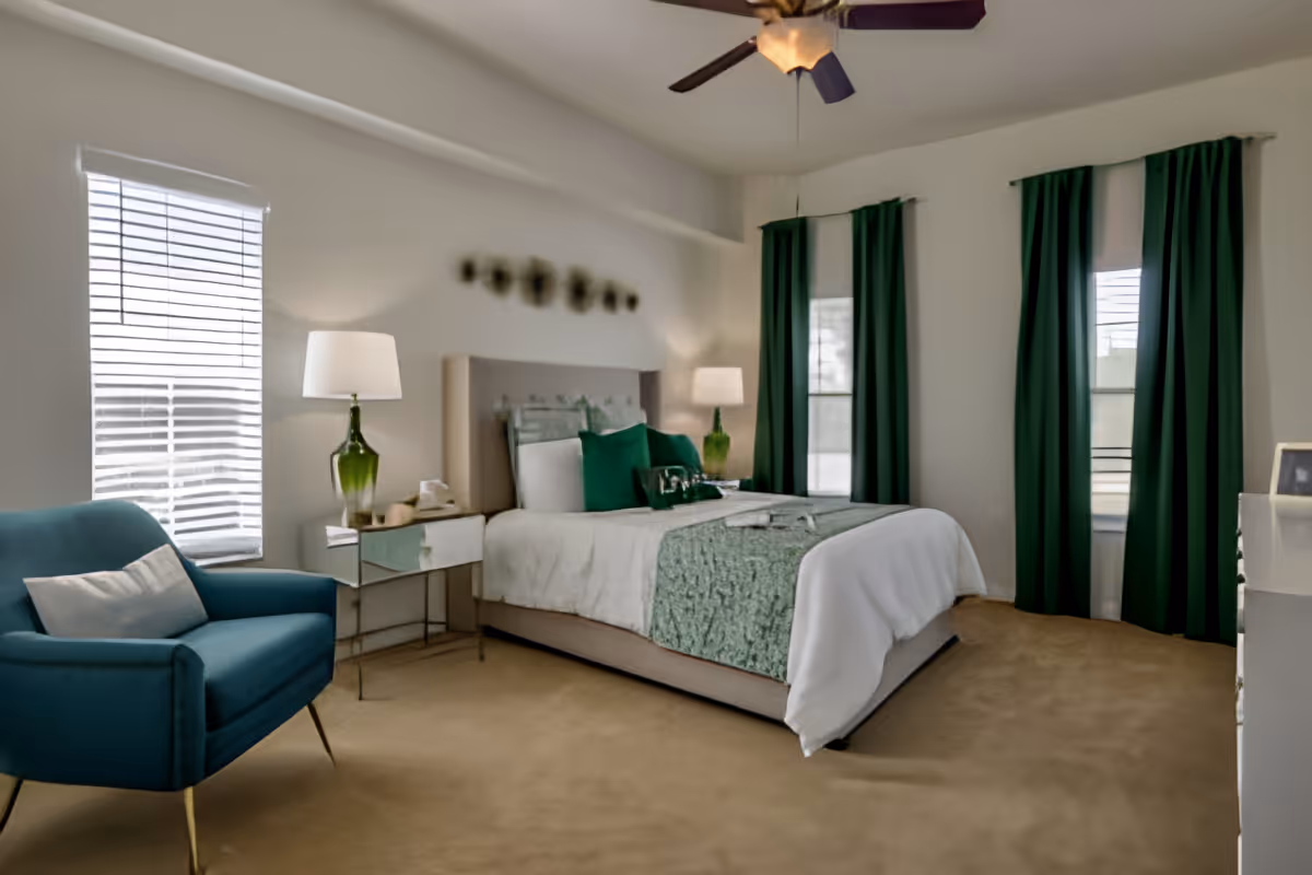 A bedroom with a large bed featuring white and green bedding, two green pillows, and a green throw blanket. There are two windows with green curtains and white blinds. A blue armchair with a white pillow is positioned near one window. Two bedside tables with green glass lamps flank the bed. The room has beige carpet and a ceiling fan with a light.