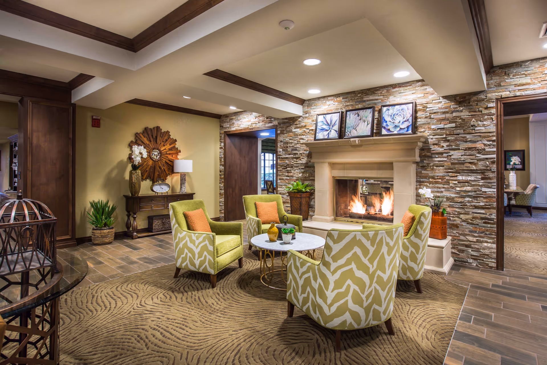 A cozy living room area with a stone fireplace lit with a warm fire. Four green patterned armchairs with orange cushions are arranged around a small round white table. The room features a textured carpet, wooden ceiling beams, and a stone accent wall with three framed floral artworks above the fireplace. There are decorative plants and a wooden console table with a lamp and clock against the wall.
