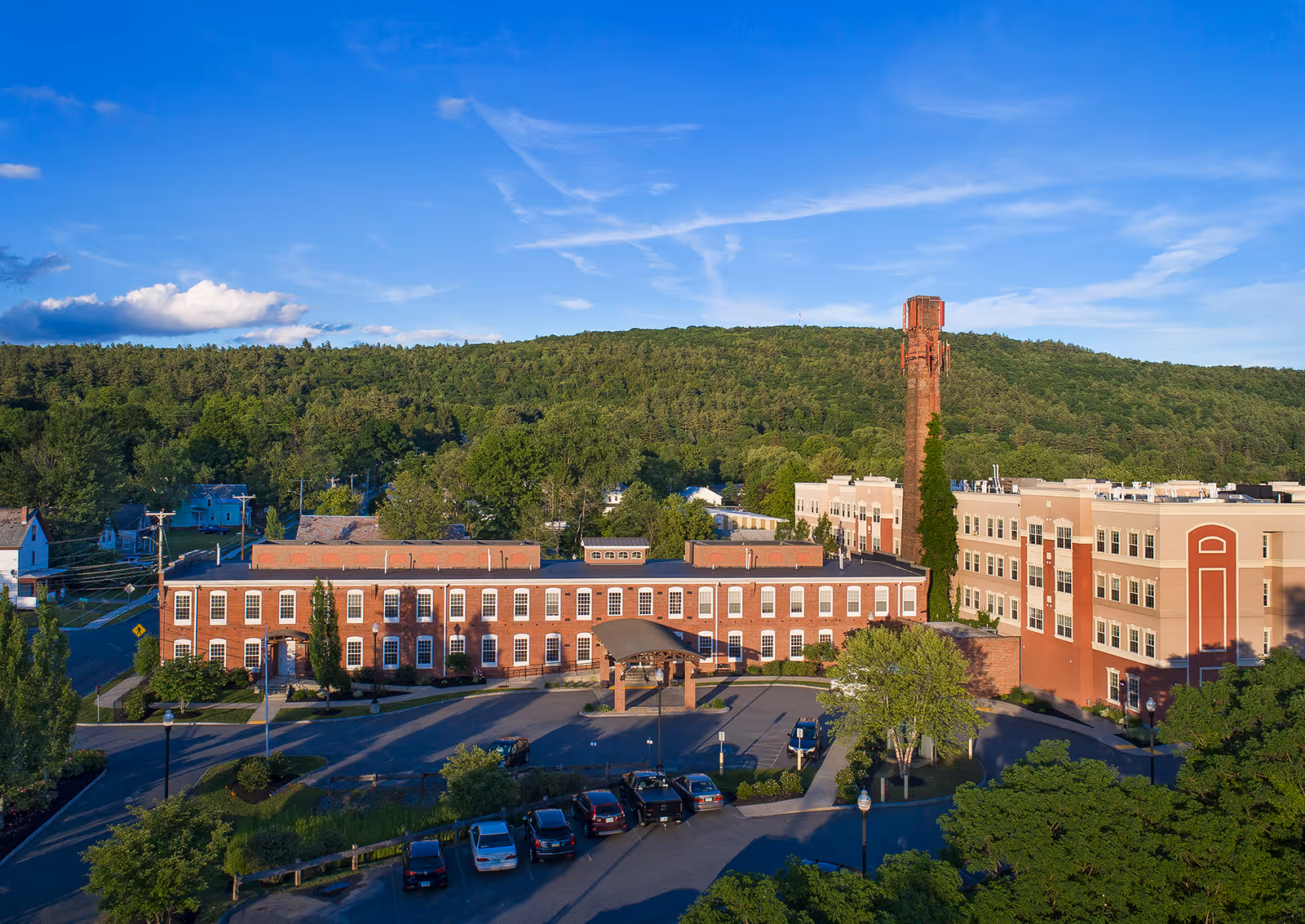 Aerial view of the American House Keene facility showing a large brick building with multiple windows, a tall chimney covered partially with ivy, a parking lot with several cars, and surrounding greenery with trees and hills in the background under a blue sky with some clouds.