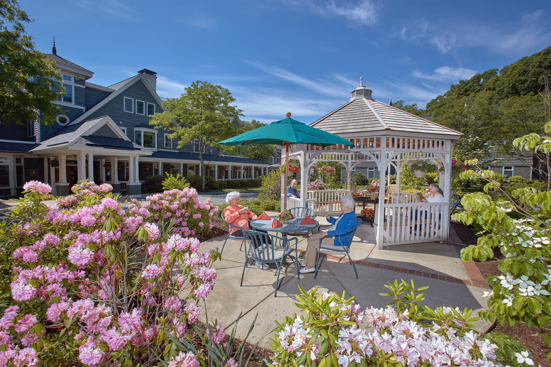 Outdoor garden area at Goddard House Assisted Living with blooming pink and white flowers, a white gazebo, and elderly residents sitting at tables under a green umbrella and inside the gazebo on a sunny day.