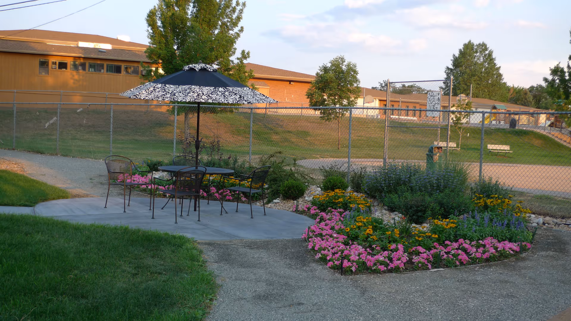 Outdoor seating area with a round table and four metal chairs under a large black and white patterned umbrella, surrounded by a garden bed with pink, yellow, and purple flowers. In the background, there is a chain-link fence, grassy areas, trees, and a building under a partly cloudy sky.