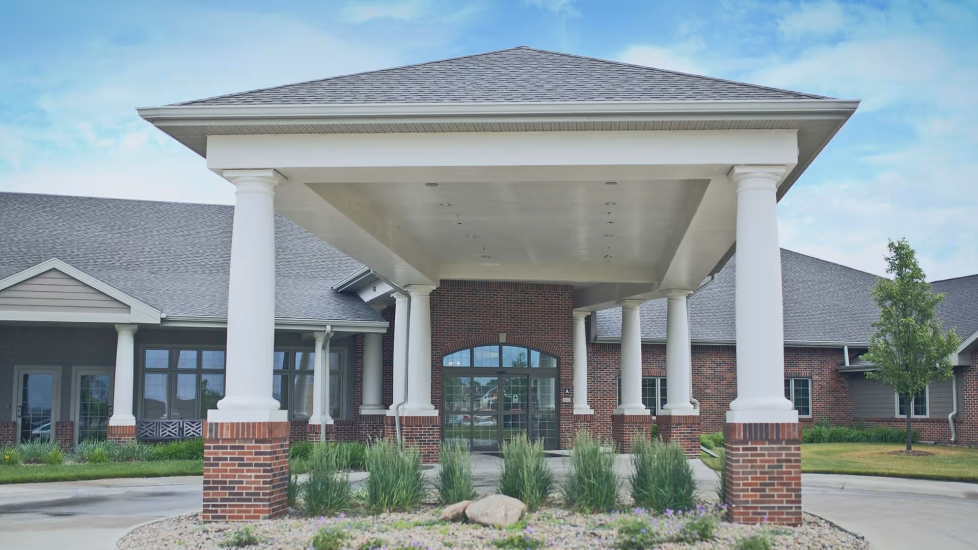 Front exterior view of Nye Square facility showing a covered entrance with large white columns on brick bases, a driveway, landscaped greenery, and a clear blue sky.