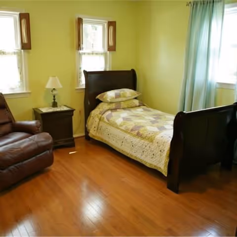 A small bedroom with a single wooden bed covered with a patchwork quilt and two pillows. There is a wooden nightstand with a lamp next to the bed, two windows with wooden shutters, a brown leather armchair, and light green walls with hardwood flooring.