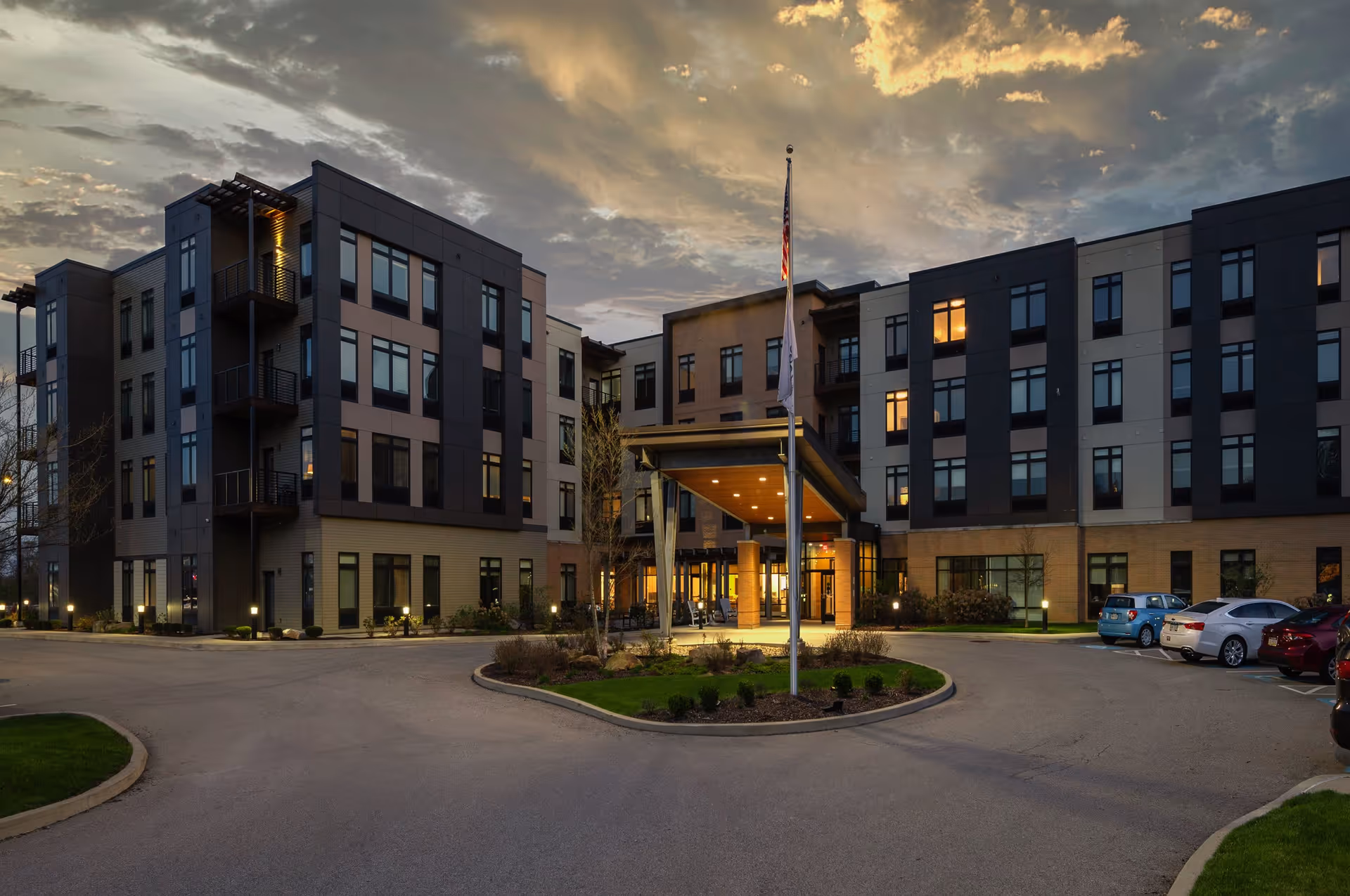 Exterior view of a modern multi-story senior living facility building at dusk with lights on inside. The entrance is covered with a canopy and there is a flagpole with an American flag in front. Several cars are parked in the parking lot to the right.