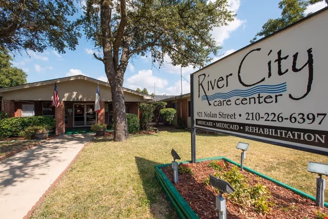 Exterior view of River City Care Center building with a large sign displaying the facility name, address, phone number, and services offered. The building is single-story with brick and siding, surrounded by grass, bushes, and a large tree. An American flag is visible near the entrance.