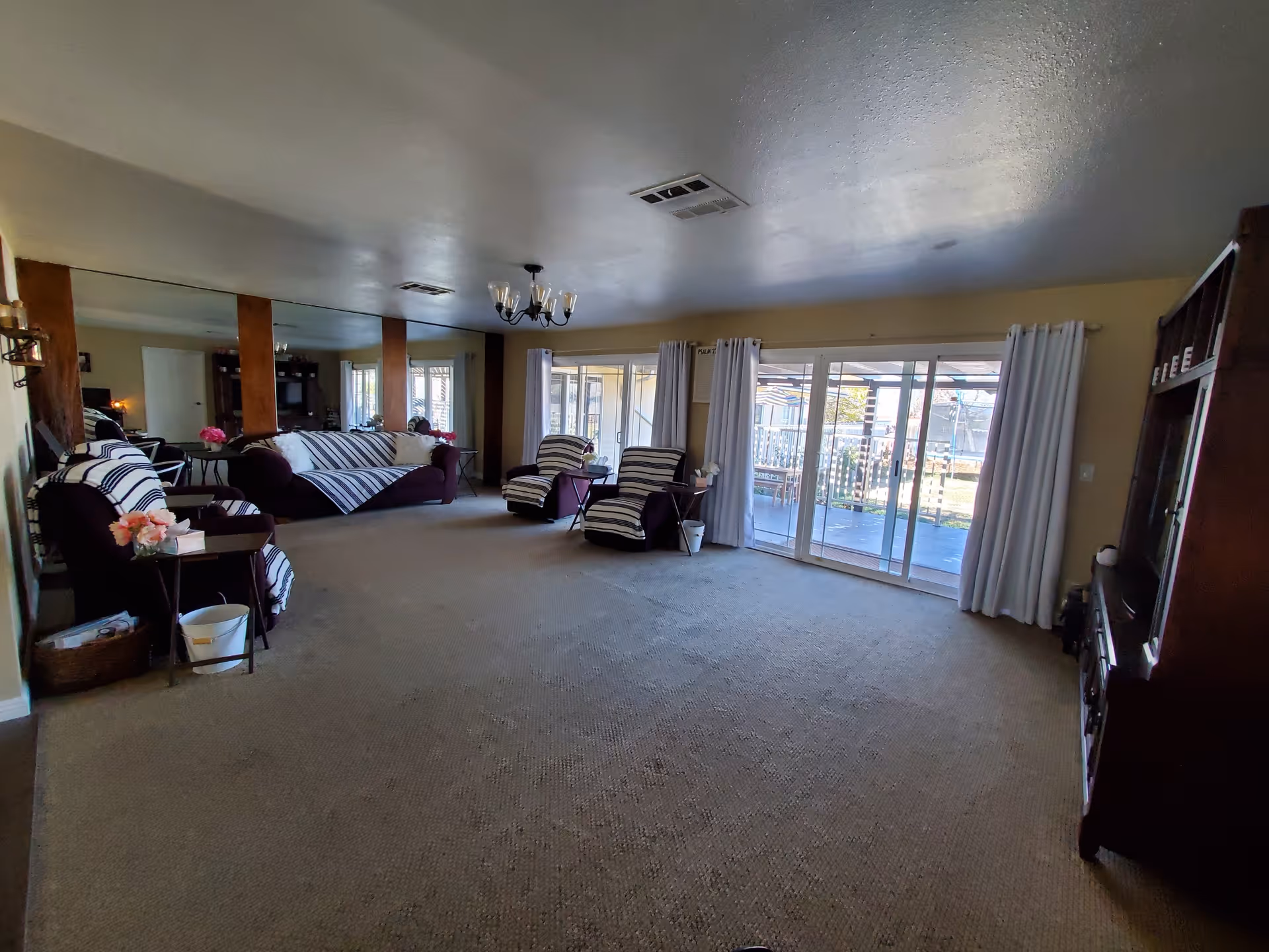 A spacious living room with beige carpet and light yellow walls. The room features a large mirror on one wall with wooden pillars, a chandelier hanging from the ceiling, and several pieces of furniture including a sofa and armchairs covered with striped fabric. There are side tables with flower arrangements and a large sliding glass door with white curtains leading to an outdoor patio area.
