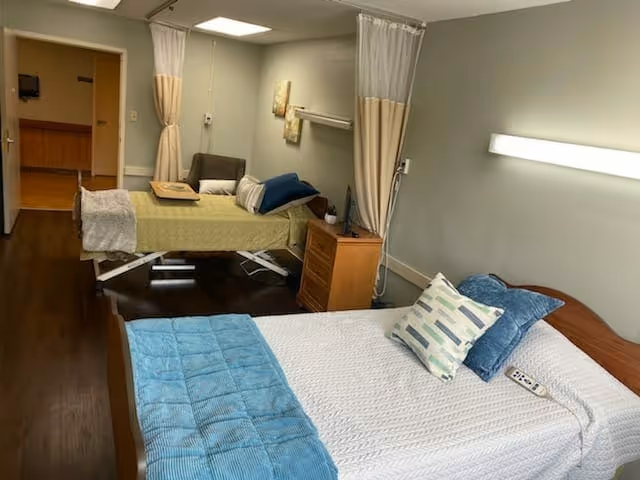 Two-bed resident room with a regular bed in front, a medical bed behind a curtain divider, a nightstand and chair.