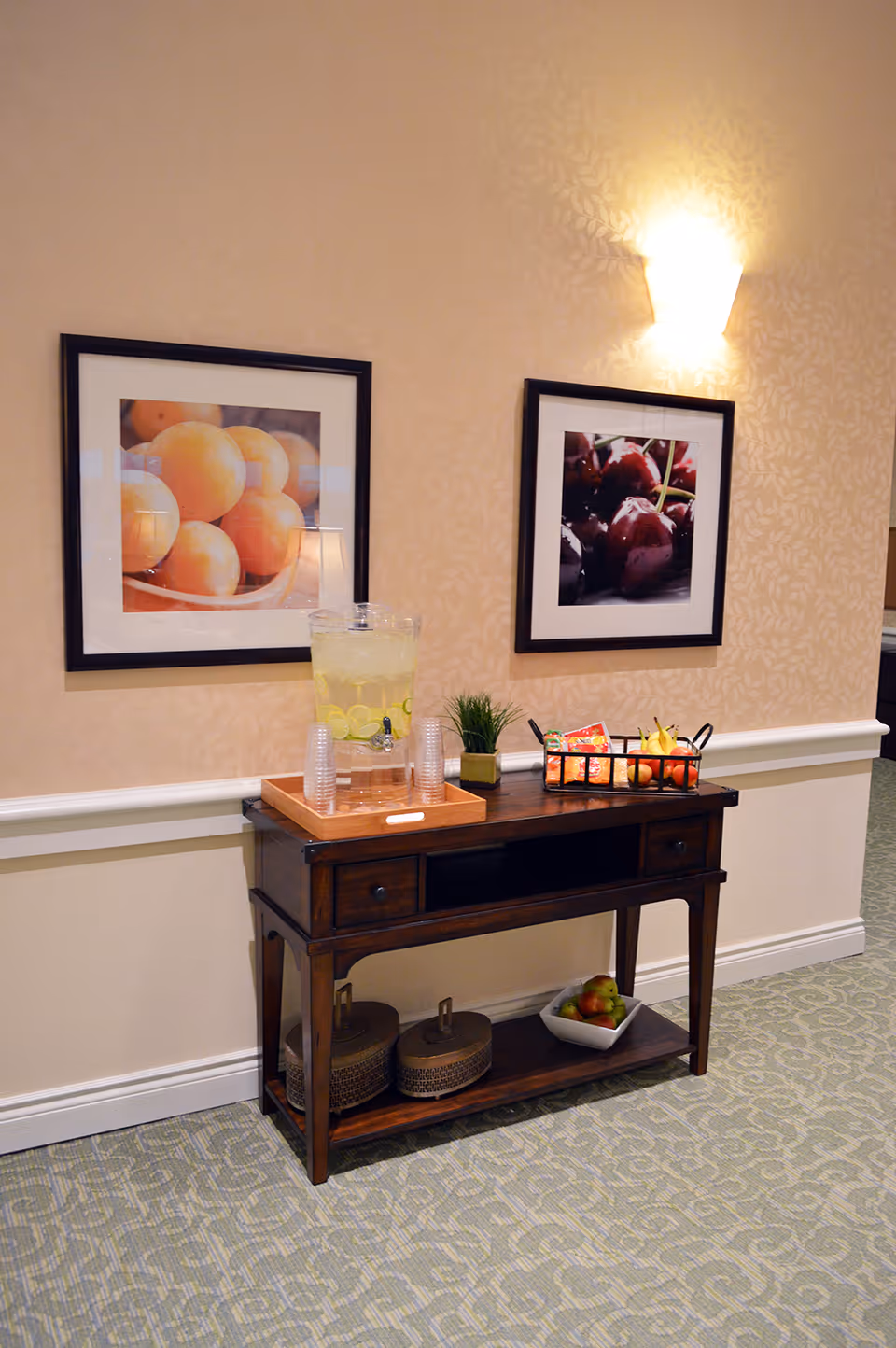 A hallway console table holding a lemon water dispenser, cups, a fruit basket and decor beneath two framed fruit prints on a patterned wall.