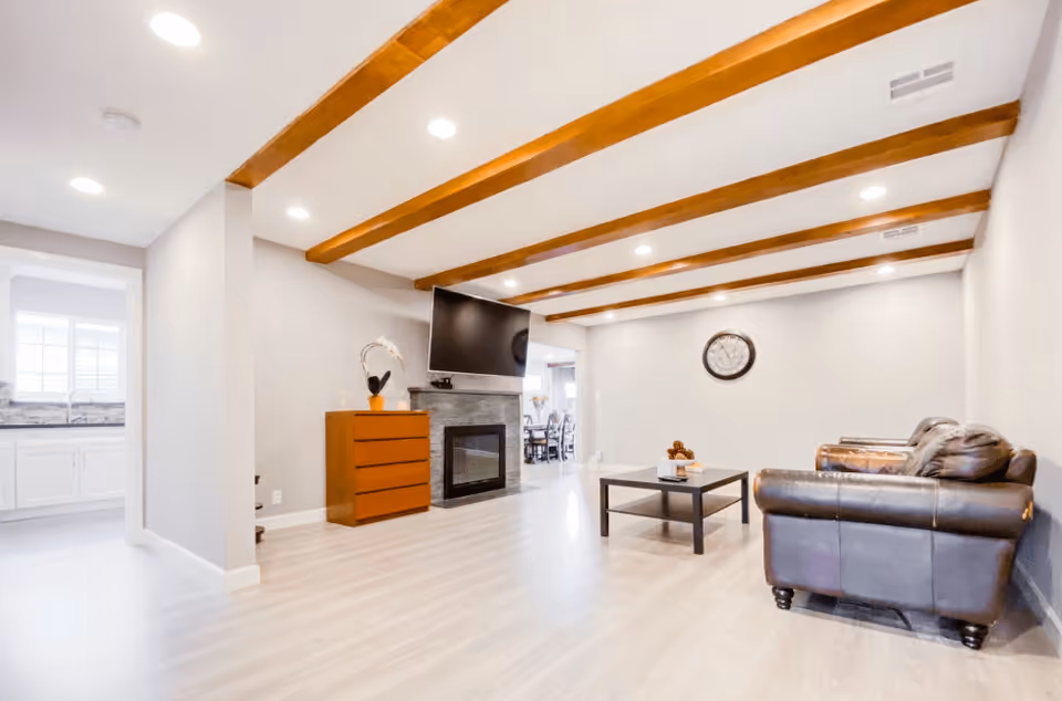A spacious living room with light wood flooring, exposed wooden ceiling beams, a wall-mounted flat-screen TV above a modern fireplace, a brown leather sofa, a black coffee table with decorative items, and a wooden chest of drawers with a plant on top. The room is well-lit with recessed ceiling lights and has a large wall clock. A kitchen area with white cabinets and a window is visible through an open doorway.
