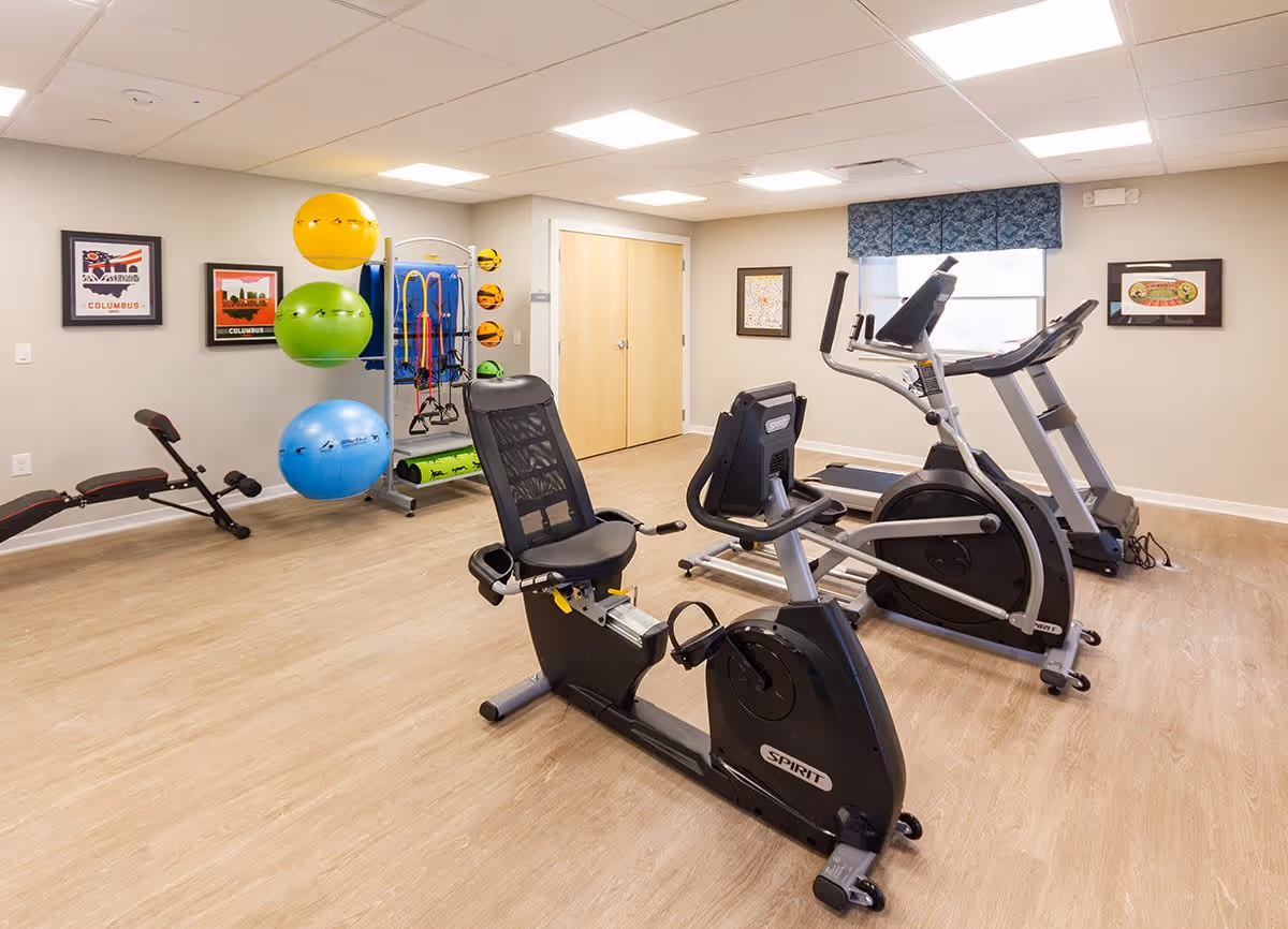 Interior exercise room with a recumbent bike, elliptical machines, a workout bench, and colorful stability balls on a rack.