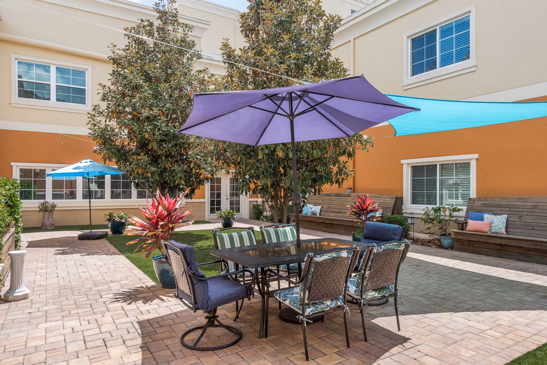 Outdoor courtyard area with a table and six chairs under a large purple umbrella. There are two large trees, potted plants, and two wooden benches with cushions along the walls of a two-story building with beige and orange walls and multiple windows.