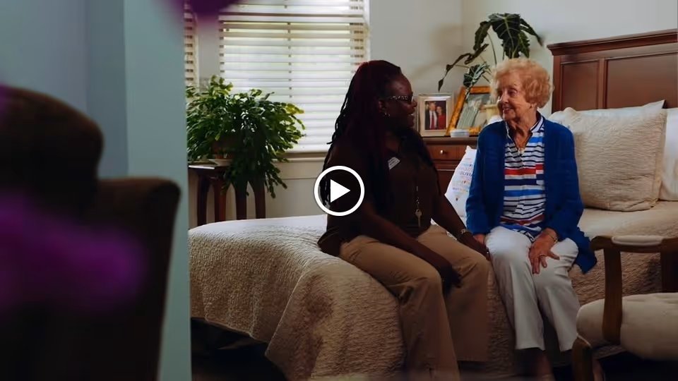 An elderly woman and a caregiver sitting on a bed in a warmly lit bedroom, engaged in conversation. The room features a large bed with a beige quilt, a wooden dresser with framed photos, a green potted plant, and a window with blinds letting in natural light.
