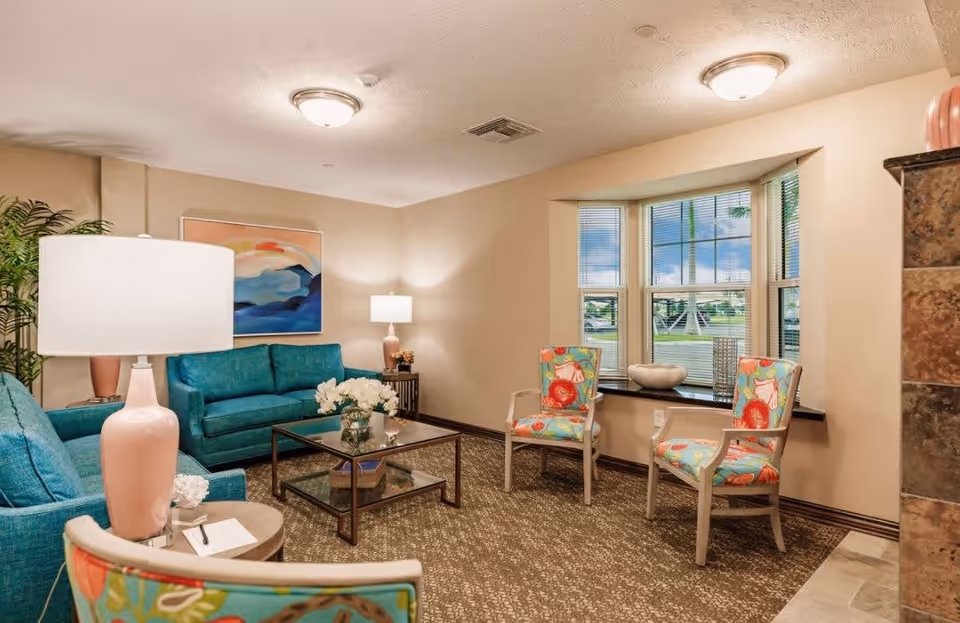 A cozy living room in a retirement community featuring teal sofas, two floral-patterned armchairs, a glass coffee table with a flower vase, a side table with a pink lamp, a large window with blinds, and a colorful abstract painting on the wall.