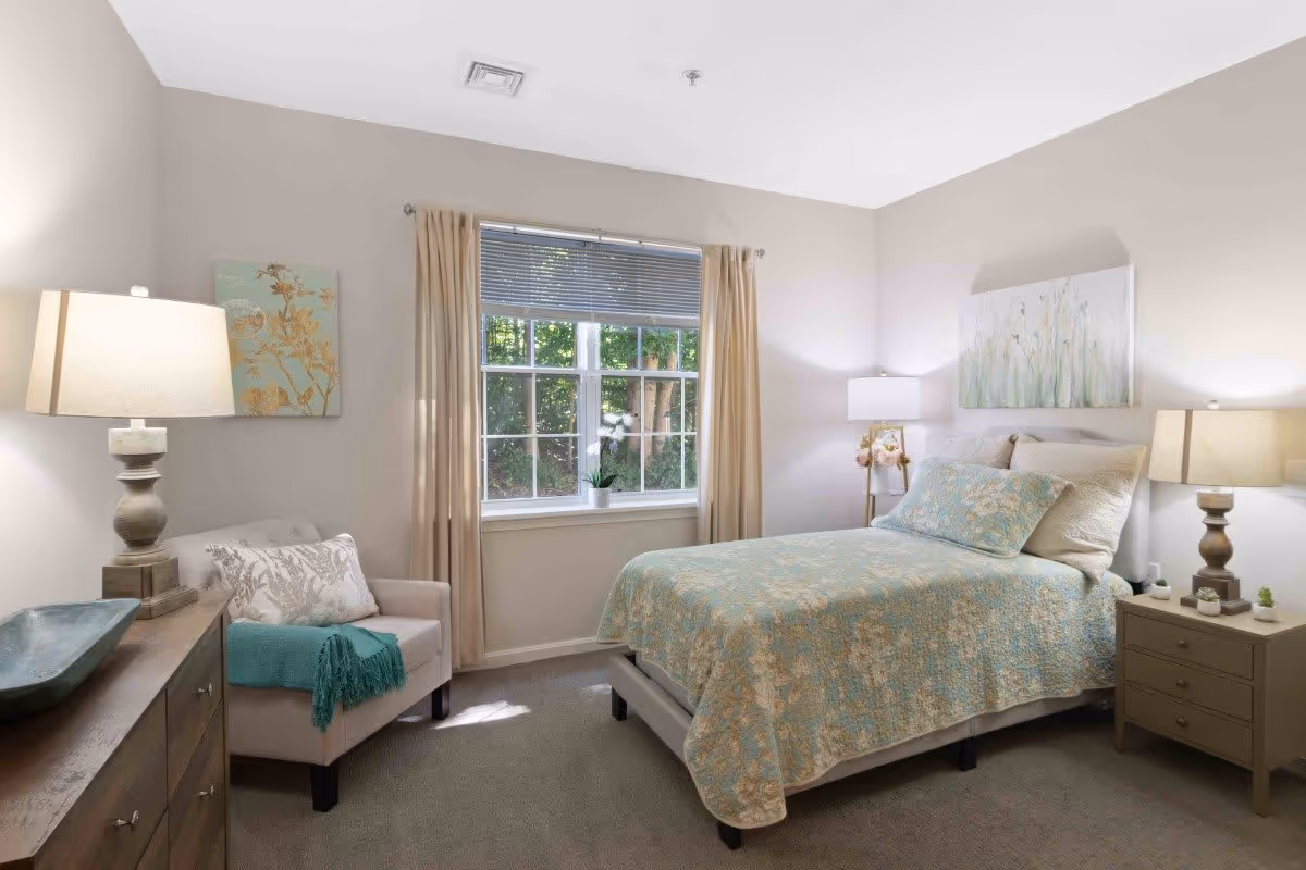 A cozy bedroom with a bed covered in a light blue and beige floral quilt, two pillows, and a painting above the headboard. There are two bedside tables with lamps on each side of the bed. A window with beige curtains lets in natural light, and a comfortable armchair with a pillow and teal throw blanket is positioned next to a wooden dresser with a lamp and decorative bowl.