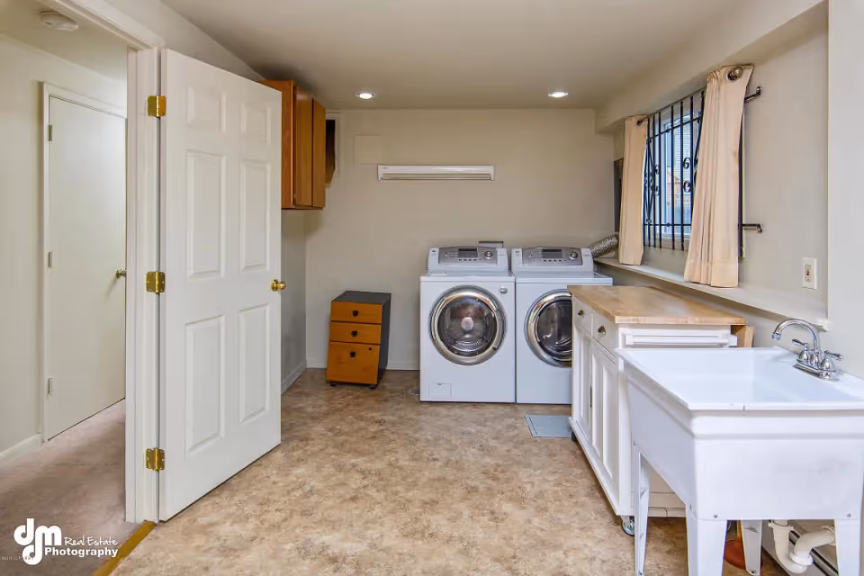 Laundry room with a white washing machine and dryer side by side, a utility sink with a faucet, a wooden countertop, a window with beige curtains and metal bars, and an open door leading to another room.