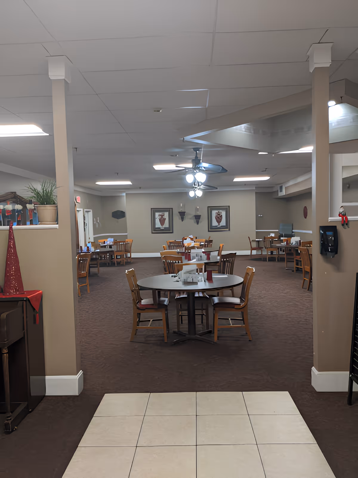 Interior view of a dining room in a senior living facility with multiple wooden tables and chairs arranged neatly. The room has beige walls, carpeted floor, ceiling fans with lights, framed artwork on the walls, and a tiled entryway in the foreground.