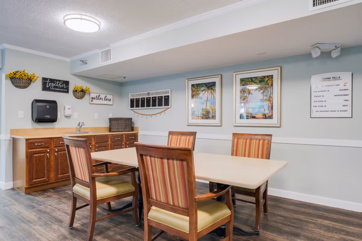 A small dining area in a senior living facility with a rectangular table surrounded by four wooden chairs with striped cushions. The room has light blue walls with white trim, two framed tropical beach paintings, and a bulletin board with the date and weather information. There is a countertop with wooden cabinets, a sink, a paper towel dispenser, and decorative wall signs that say 'together is my favorite place' and 'gather here'.