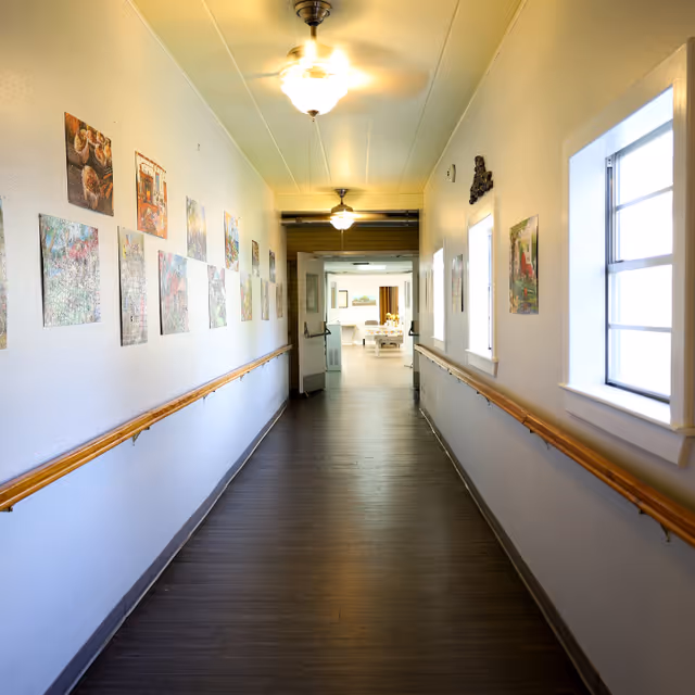A well-lit hallway in a senior living facility with wooden handrails on both sides, decorated with various framed pictures on the left wall and windows on the right. Ceiling fans with lights are mounted on the ceiling, and the hallway leads to a bright room with tables and chairs visible in the distance.
