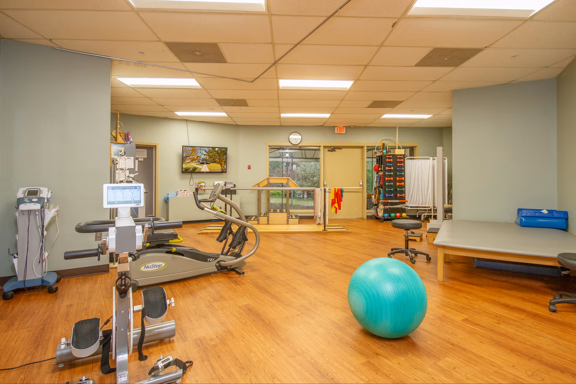 Interior rehab/therapy room with exercise equipment, a large turquoise exercise ball, weights, and a treatment table on wood flooring.
