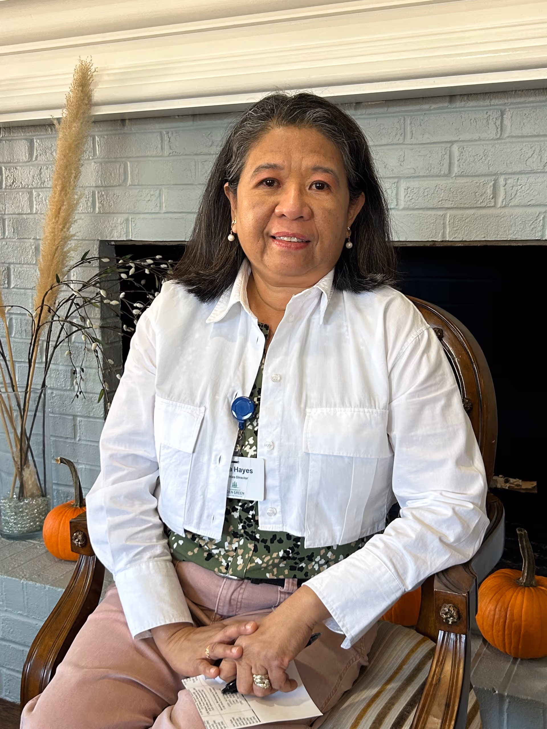 A woman with gray hair wearing a white cropped jacket and a patterned green blouse sits on a wooden chair in front of a gray brick fireplace. There are two small pumpkins and decorative dried plants on the hearth behind her.