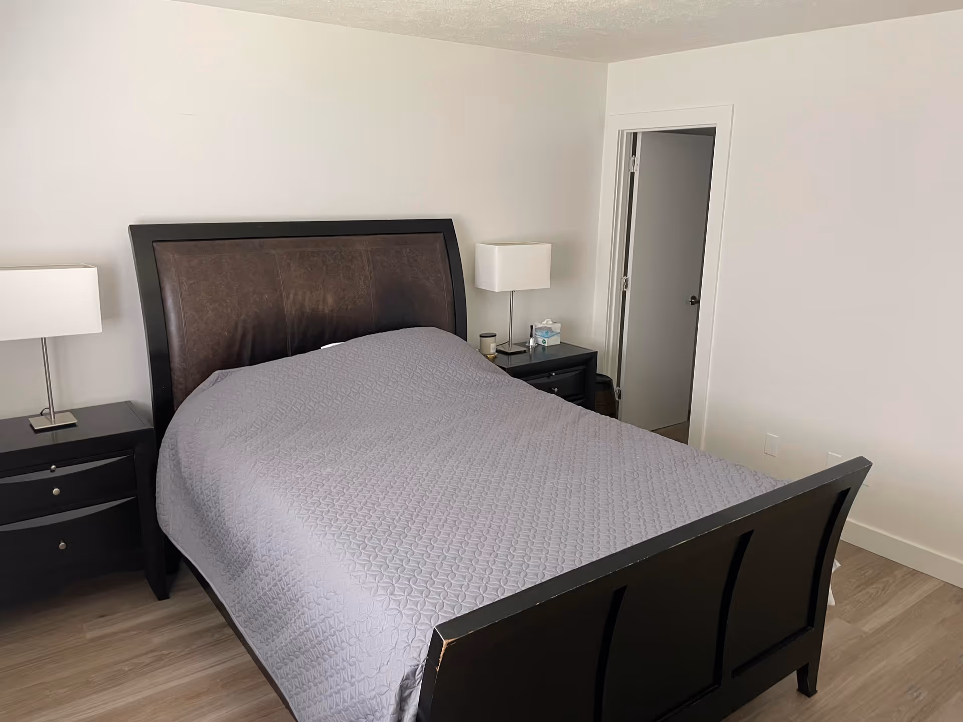 A bedroom with a dark wooden bed frame and a gray quilted bedspread. There are two matching dark wooden nightstands on either side of the bed, each with a modern white lamp. The room has light-colored walls and wooden flooring. A doorway is visible on the right side of the image.