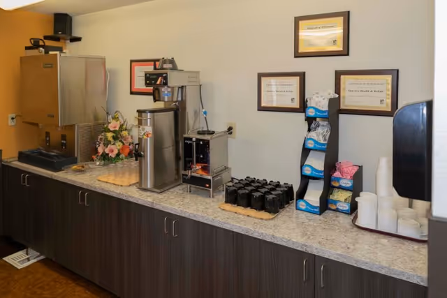 A countertop with various beverage and snack dispensers including a coffee machine, a hot water dispenser, a small toaster oven, and a water dispenser. There are black mugs, disposable cups, napkins, and sweetener packets arranged neatly on the counter. Three framed certificates hang on the wall above the counter.