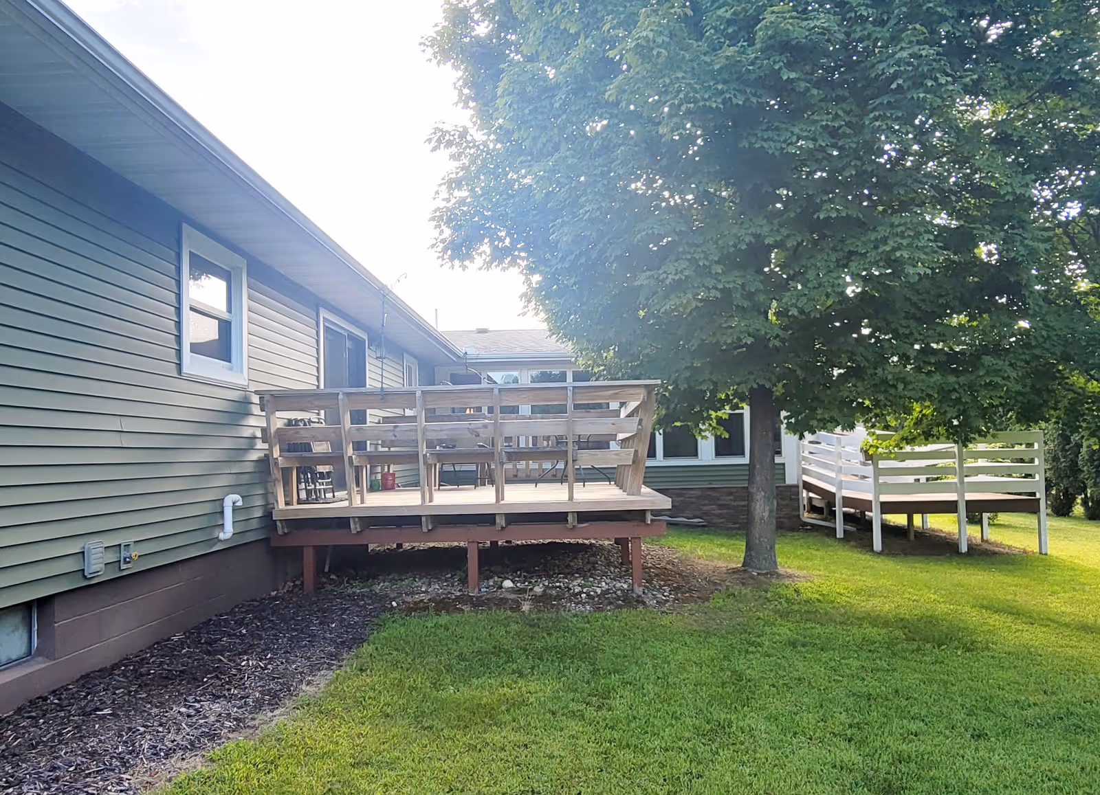Outdoor view of a single-story building with green siding and a small wooden deck attached. There is a large leafy tree next to the deck and a white wooden ramp leading to another entrance. The area is surrounded by green grass and landscaping.