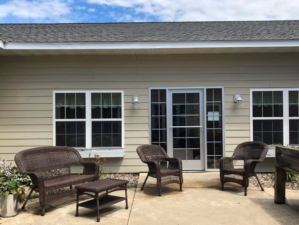 Outdoor patio area with three brown wicker chairs and a matching wicker coffee table on a concrete surface. The patio is in front of a beige building with two windows on either side of a glass door. There are small plants and rocks along the building's foundation.