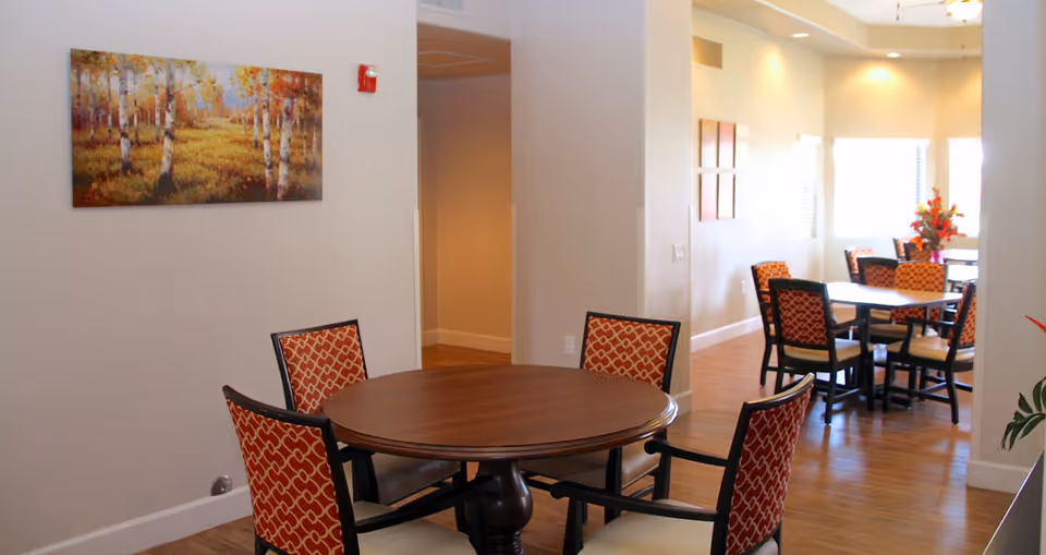 Bright communal dining area with round wooden tables and patterned upholstered chairs, wall art, and windows letting in light.