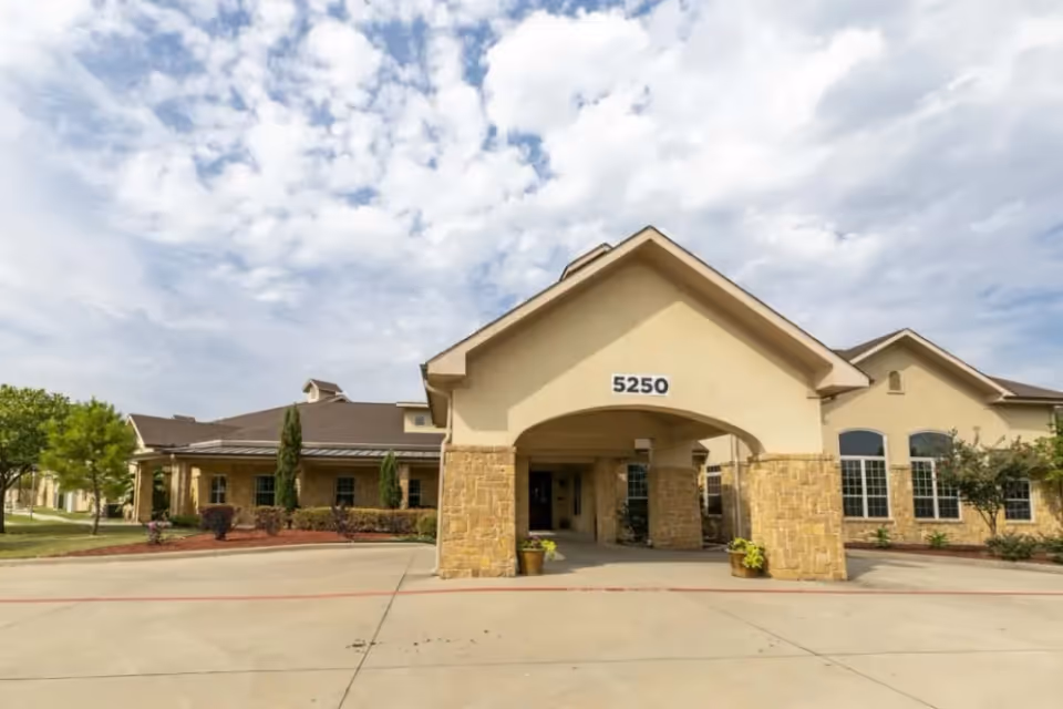 Front exterior view of a single-story assisted living facility building with a covered entrance supported by stone pillars, the number 5250 displayed above the entrance, surrounded by landscaped greenery and a partly cloudy sky.