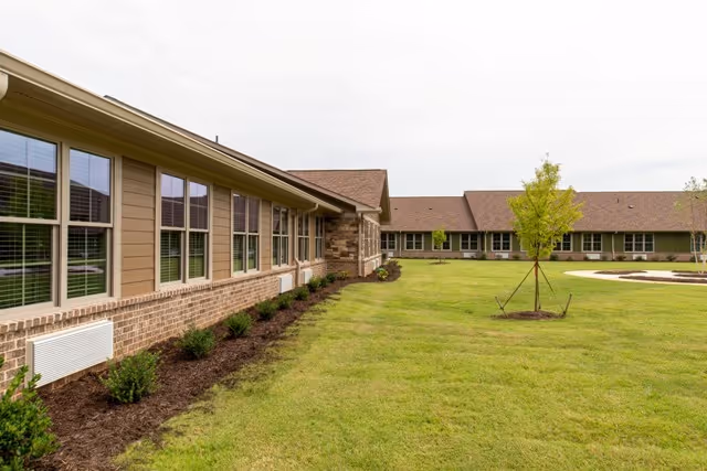 Exterior view of a single-story assisted living facility building with multiple windows, a well-maintained lawn, small shrubs along the building, and a young tree supported by stakes in the grassy area.