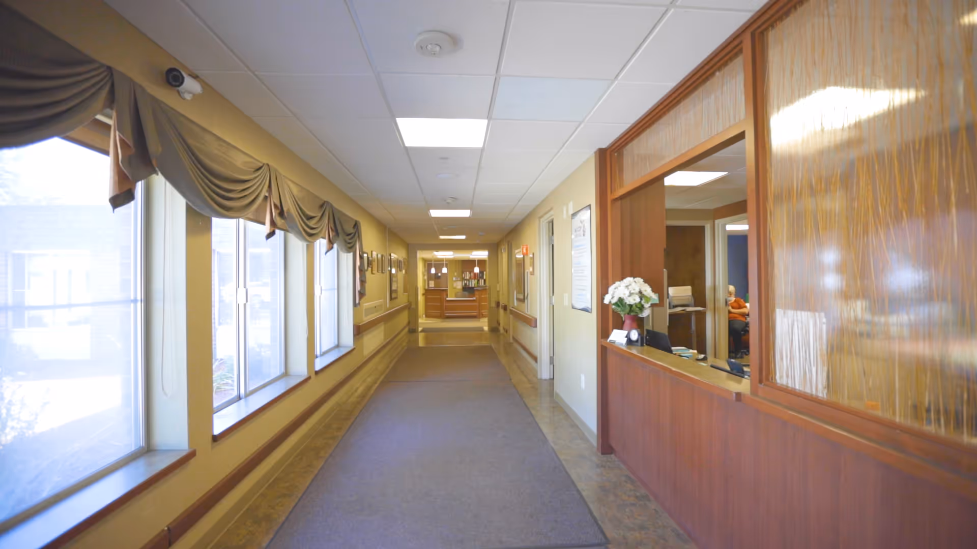 A well-lit hallway in a senior living facility with large windows on the left side covered by draped valances. On the right side, there is a wooden reception desk with a vase of white flowers and a person sitting behind it. The hallway has a carpet runner and leads to another room with more lighting fixtures visible.